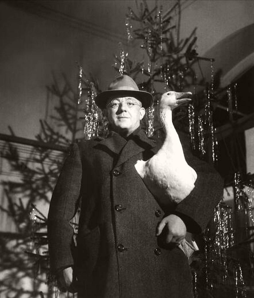 Photograph of British actor, Alec Guinness, standing before a Christmas tree while clutching a goose