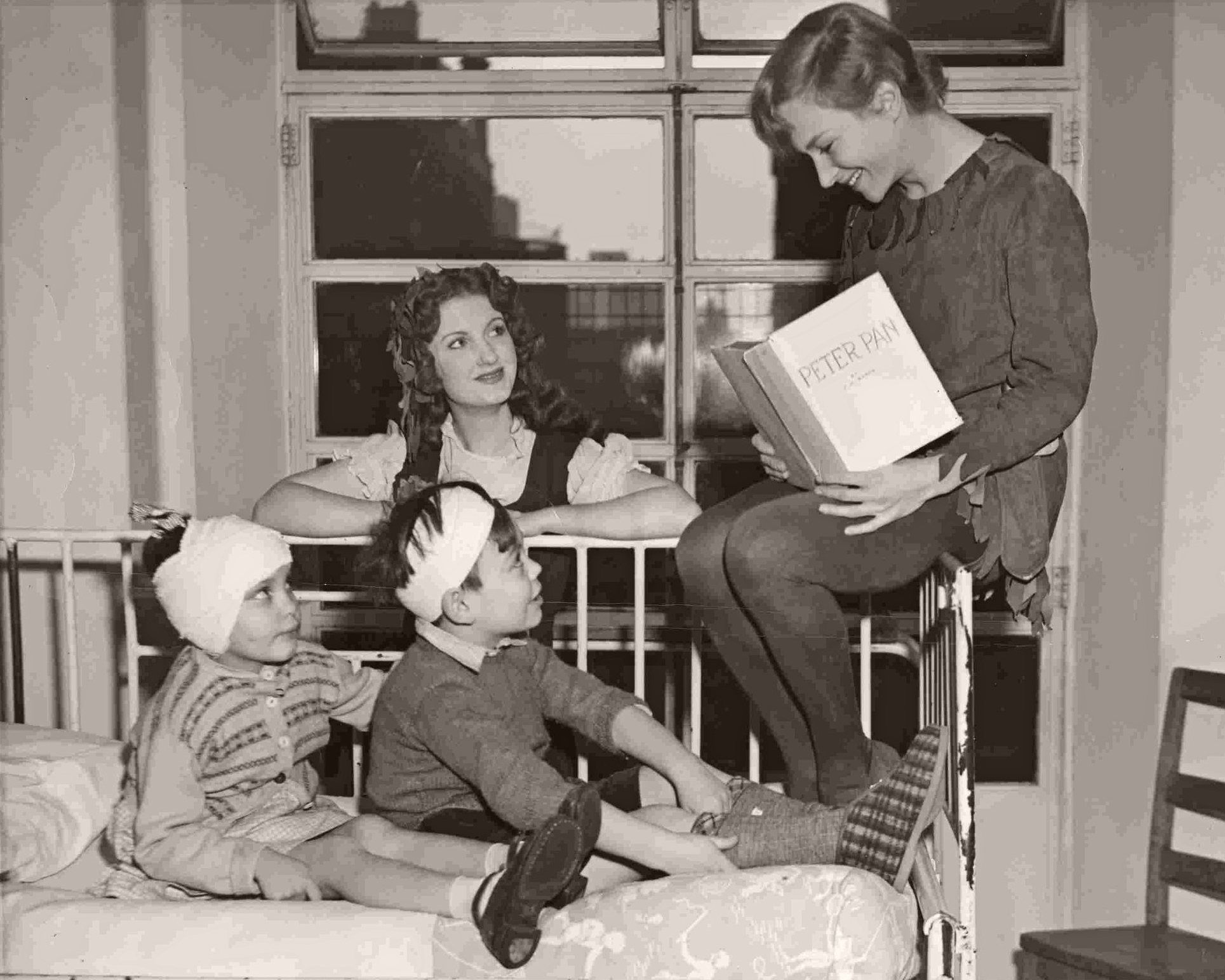 British actress Joan Greenwood, in costume as Peter Pan, sits on the end of a ward bed and reads to the sick children