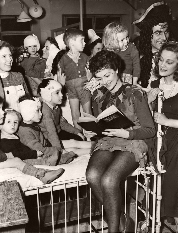 Margaret Lockwood, in costume as Peter Pan, perches on a bed in the ward as she reads to the children at the Great Ormond Street Hospital in 1950