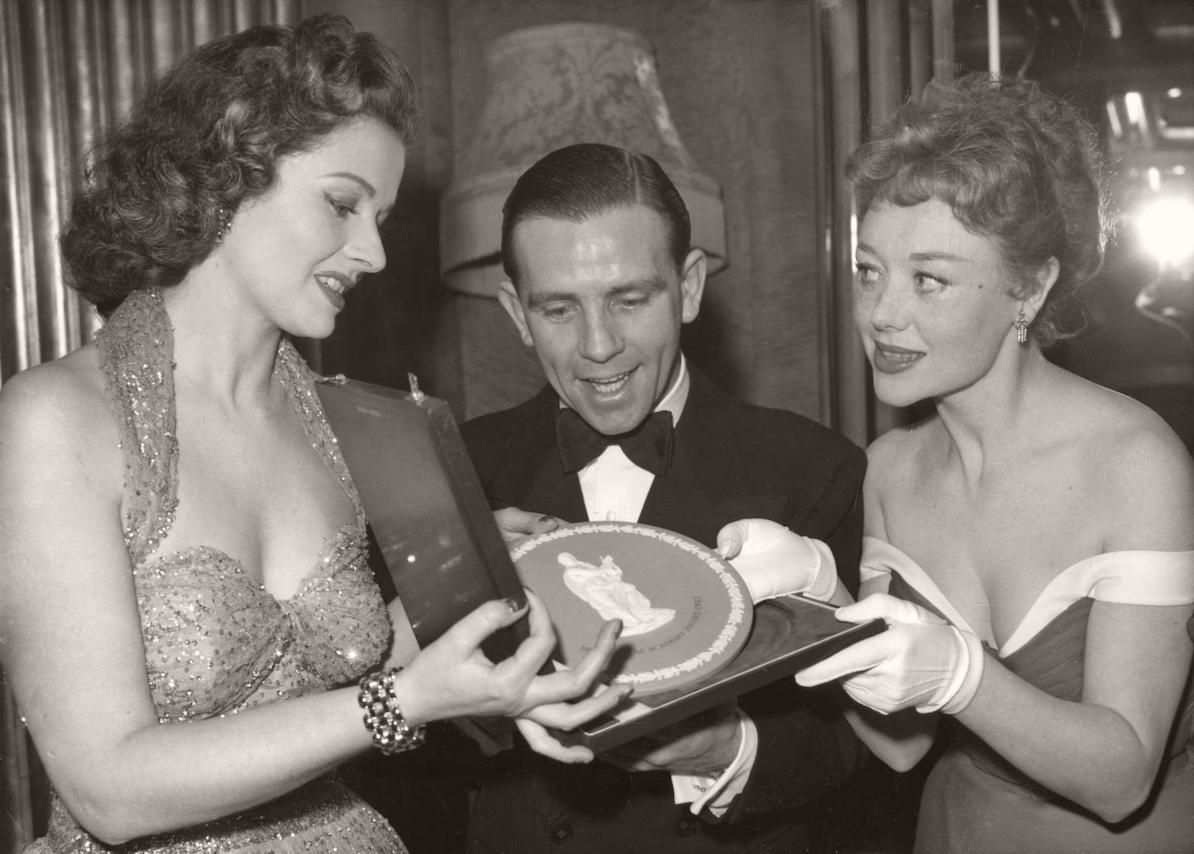 Norman Wisdom, dressed in a dinner jacket, smiles as looks at his British Film Academy [now BAFTA] award of 1953 for ‘Most Promising Newcomer to Leading Film Roles’. Actresses Margaret Lockwood and Glynis Johns look on