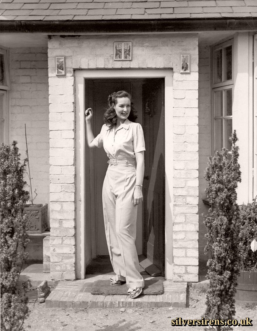 Patricia Roc stands on the steps of a brick porch.  The actress is dressed in a light-coloured trouser suit and sandals.