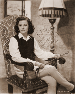 Margaret Lockwood holds a riding crop while she sits beneath a standard lamp in an armchair